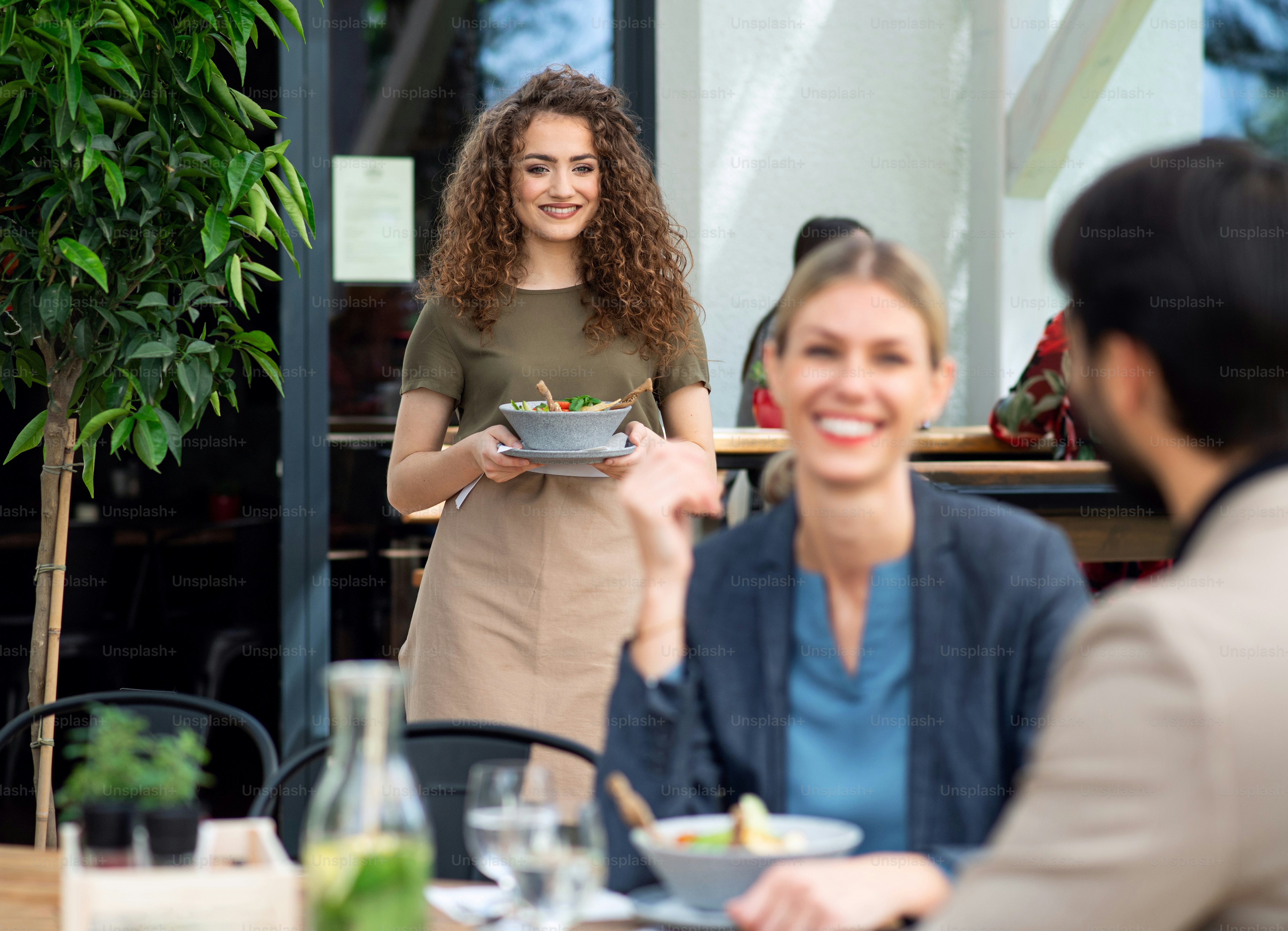Woman waitress with face mask serving happy couple outdoors on terrace ...