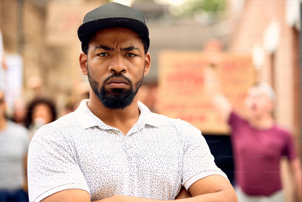 African American man standing with arms crossed in front of crowd of people during anti-racism protest.