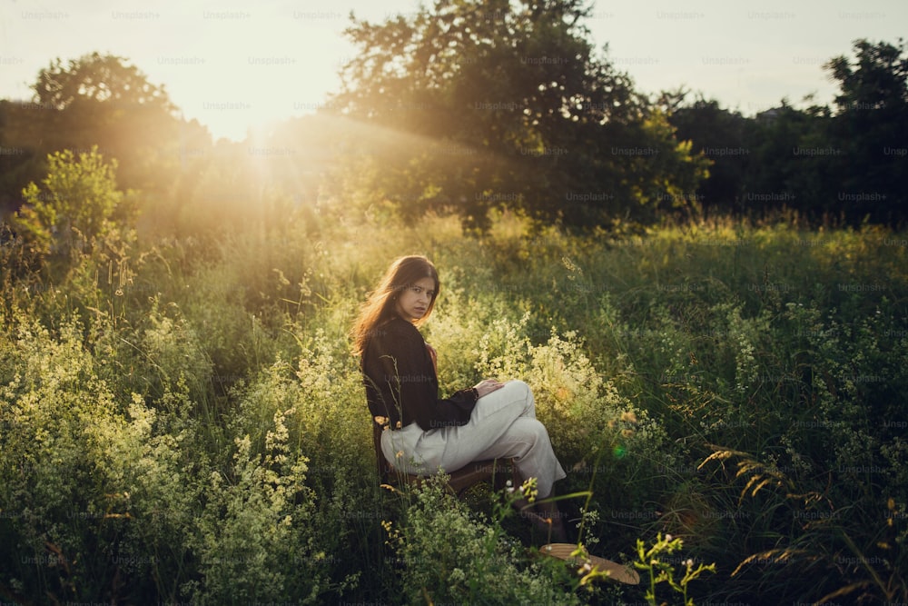 Fashionable woman relaxing in summer countryside. Stylish elegant girl sitting on rustic chair in sunset light in summer meadow.Atmospheric tranquil moment. Creative image.