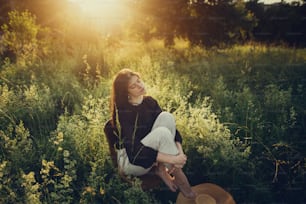 Fashionable woman relaxing in summer countryside. Stylish elegant girl sitting on rustic chair in sunset light in summer meadow.Atmospheric tranquil moment. Creative image.