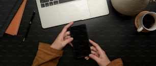 Overhead shot of hands holding smartphone on dark modern workspace with laptop, supplies and decoration