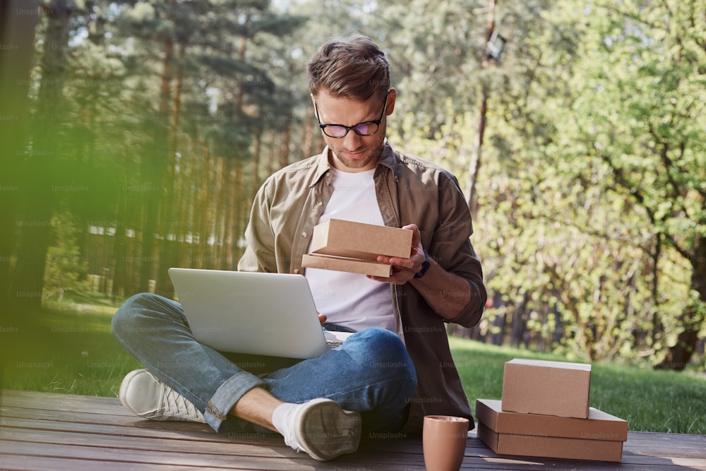 Busy stylish male scrutinizing the parcel while sitting on wooden surface with crossed legs