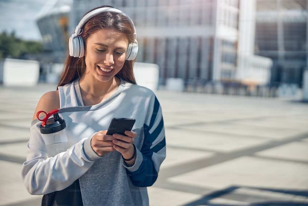 Waist-up portrait of a high-spirited sporty lady staring at her cellular phone in her hands