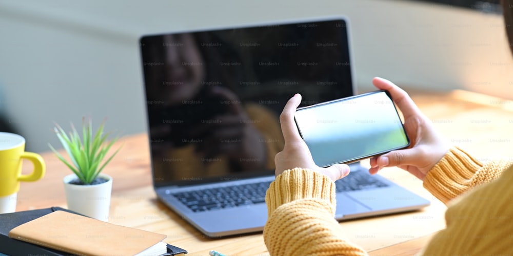 Imagem cortada da mão está usando um smartphone enquanto está sentado na frente de um laptop de computador de tela vazia.