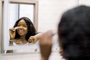 Reflection of pretty smiling African girl .mirror applying cosmetic cream on her face. Female putting moisturizer on her facial skin in light home bathroom. Spa and beauty at home