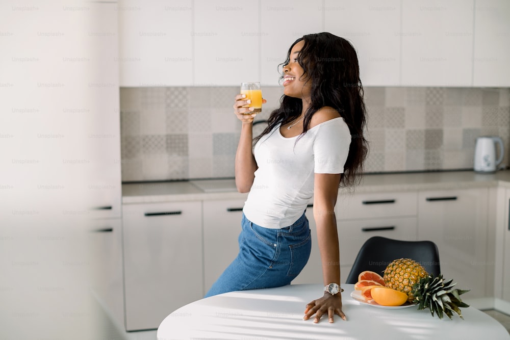 Young beautiful African American woman holding glass with an orange juice, looking at window, posing at light modern kitchen at home. Healthy lifestyle concept, natural organic breakfast at kitchen.