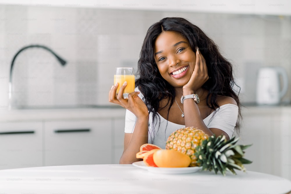 Porträt einer jungen lächelnden afrikanischen Frau, die drinnen in der gemütlichen Küche sitzt, am Tisch mit frischem Obst und hält ein Glas Saft.