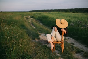 Muchacha elegante con estilo en sombrero de paja sentada en silla rústica en prado de verano en la noche. Mujer joven de moda relajándose en el campo, momento tranquilo. Imagen creativa. Campo de verano