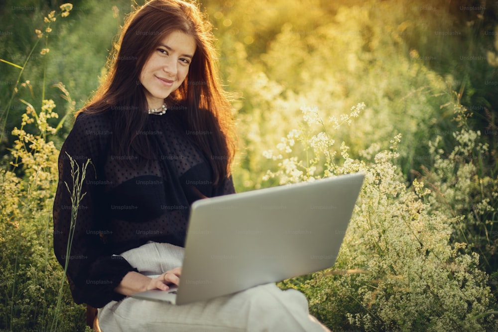 Chica elegante de moda trabajando en una laptop y sentada en una silla rústica bajo el cálido sol en el prado de verano al atardecer. Mujer de negocios joven que trabaja en línea al aire libre. Nuevo concepto de oficina