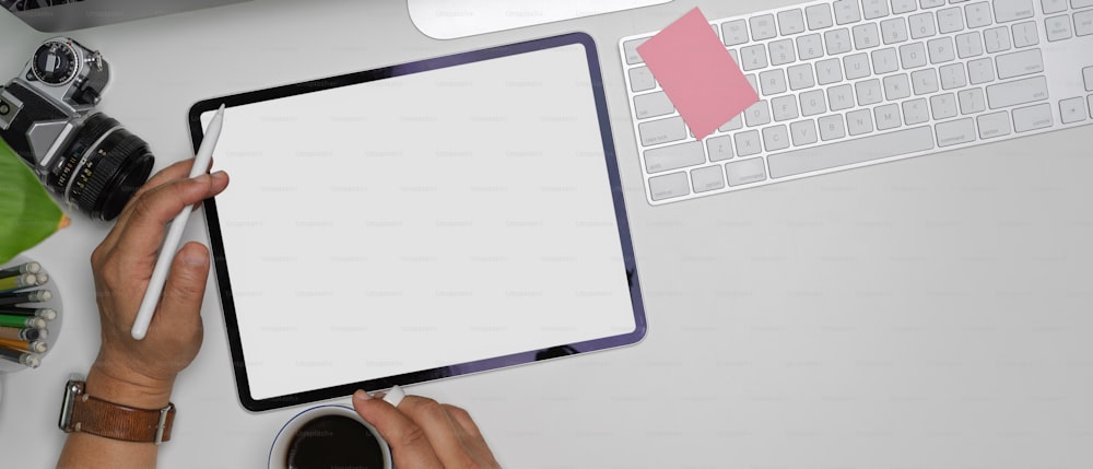 Overhead shot of male left hand using mock-up tablet while right hand holding coffee cup on white office desk