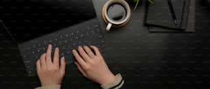 Overhead shot of hands typing on digital tablet keyboard on dark modern office desk with coffee cup and schedule books