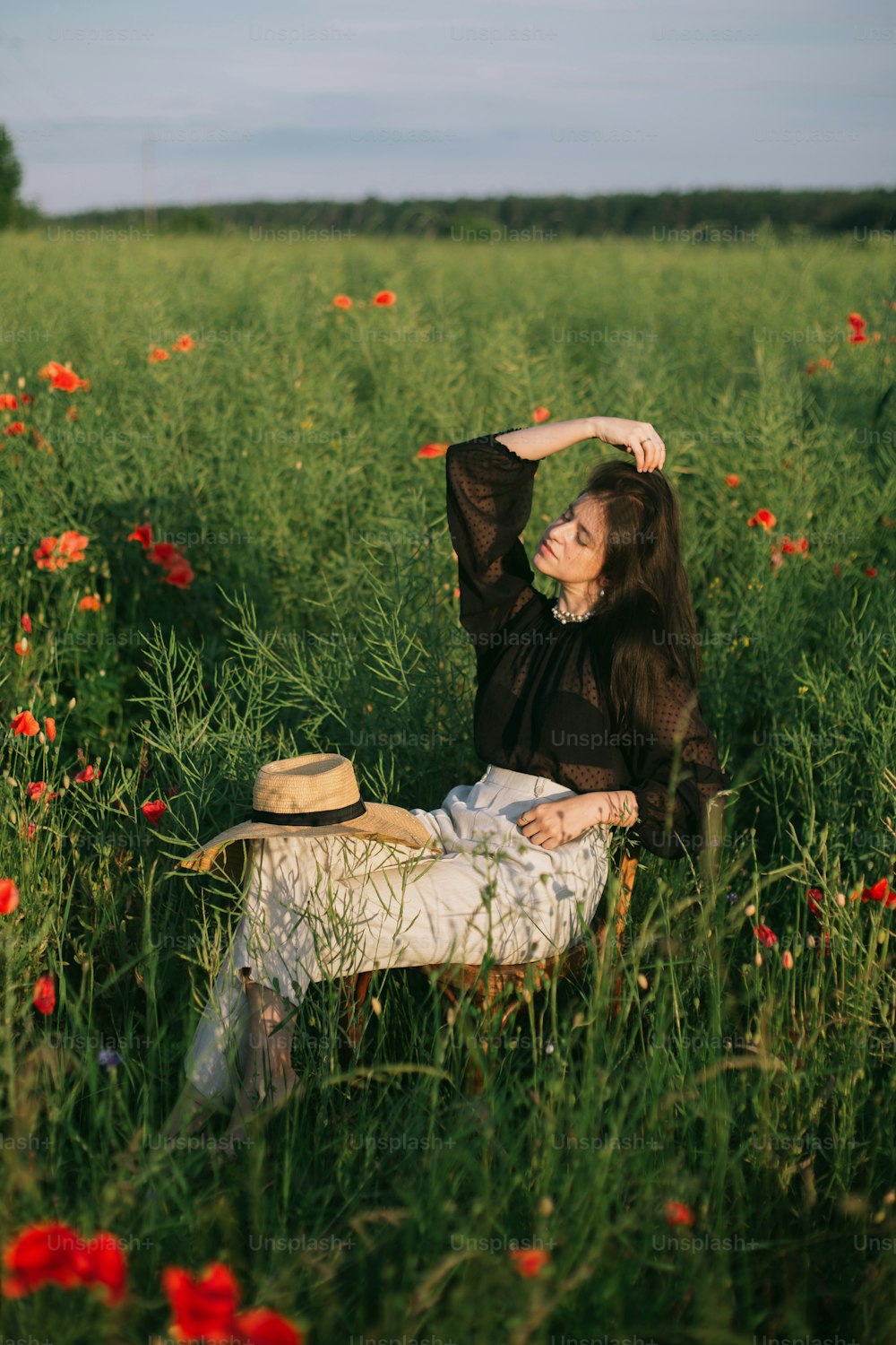 Slow living. Stylish elegant girl sitting on rustic chair in summer meadow with flowers. Fashionable young woman relaxing in field in evening light. Creative beautiful image.