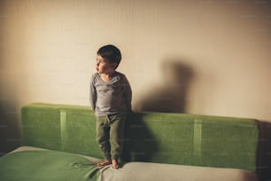 Niño caucásico soñador sentado descalzo en el sofá y mirando hacia afuera a través de la ventana durante la cuarentena