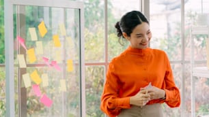 Business people work on project planning board in office and having conversation with coworker friend to analyze project development . They use sticky notes posted on glass wall to make it organized .