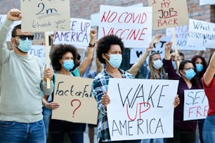 Displeased crowd of people with face masks demonstrating in city streets during coronavirus epidemic. Focus is on Black woman holding a placard with Wake up America inscription.