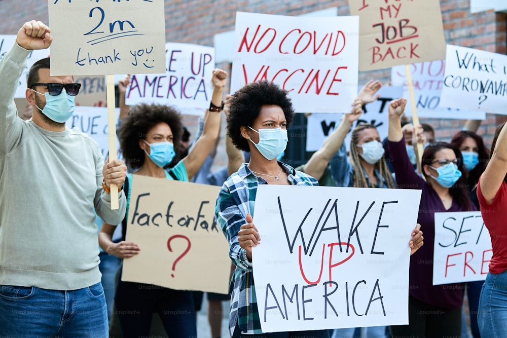 Displeased crowd of people with face masks demonstrating in city streets during coronavirus epidemic. Focus is on Black woman holding a placard with Wake up America inscription.