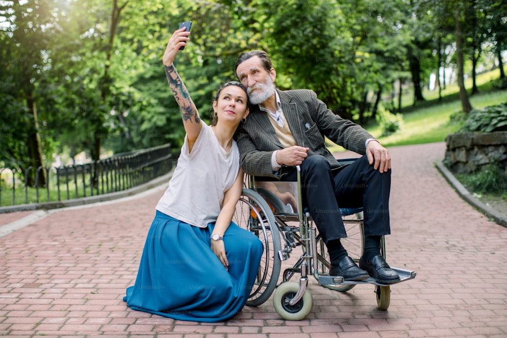 Pretty smiling young woman, care giver or granddaughter in casual hipster clothes, with dreadlock hair, making photo on smartphone with her disabled elderly man, grandfather or patient in wheelchair.
