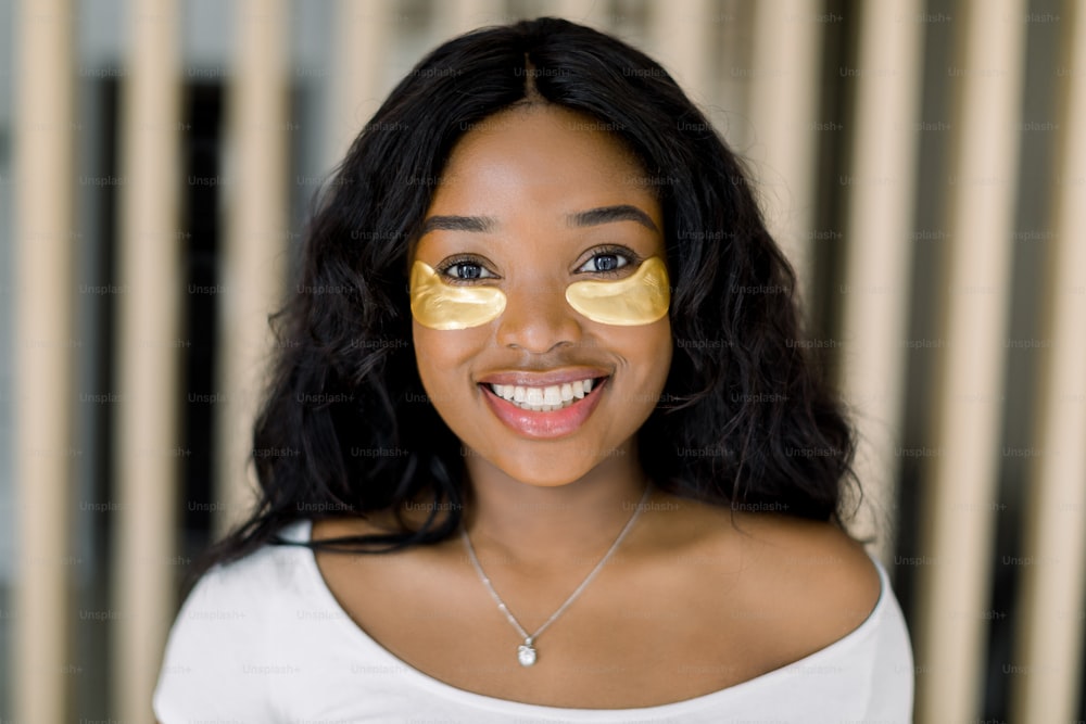 Smiling African American woman with golden under eye patches, looking at camera in home, performing daily morning facial skincare routine. Antiaging, moisturizing, beauty concept.