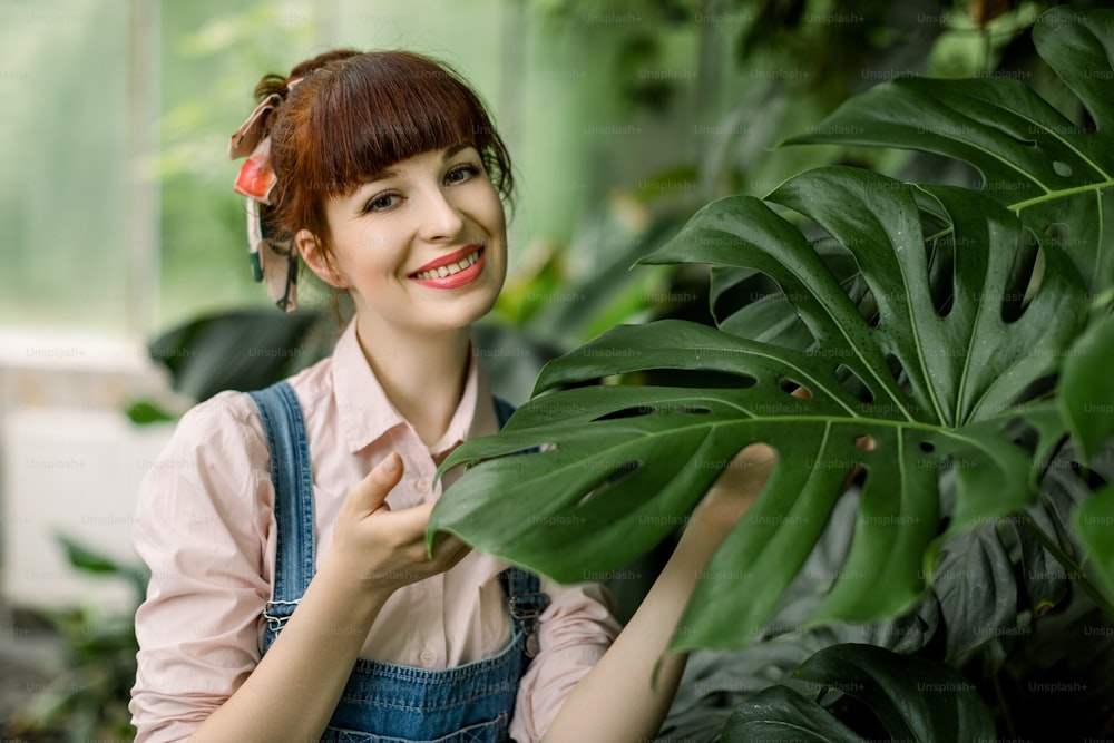 Portrait of smiling attractive red haired woman gardener, posing to camera holding monstera leaf near her face, while standing in greenhouse with plants on the background.