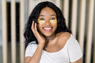 Beautiful young African woman applies gold eye mask, medical eye antiwrinkle patches. Portrait of beauty dark skinned model with natural nude make up cares about her skin. Spa, skincare at home.