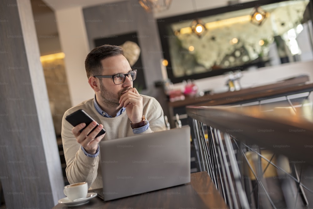 Male freelancer working remotely from a restaurant, holding a smart phone and using a laptop computer, serious and pensive