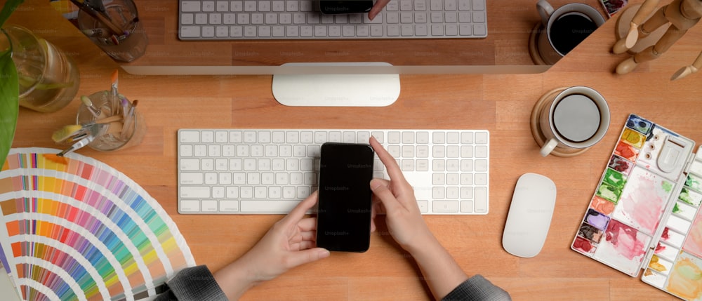 Overhead-Aufnahme einer Designerin mit Smartphone, während sie mit Computer und Malwerkzeugen am Schreibtisch sitzt