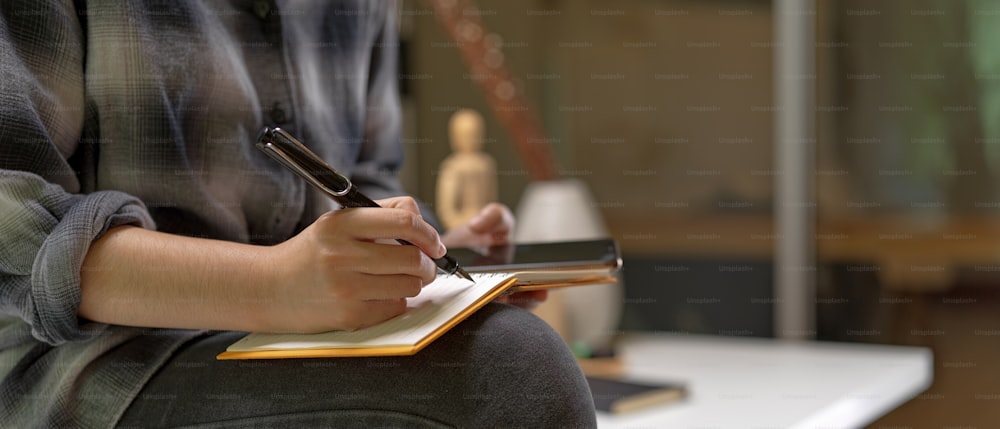 Cropped shot of female worker sitting on worktable and taking note on blank schedule book while looking on smartphone