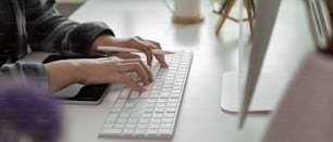 Vista lateral de manos femeninas escribiendo en el teclado de la computadora mientras sostiene el lápiz óptico en el escritorio blanco de la oficina