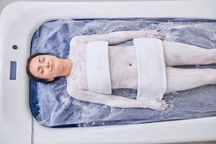 Calm young woman smiling with closed eyes while having her skin rubbed with white clay and lying on a modern spa device