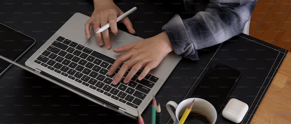 Cropped shot of hands typing on laptop keyboard on modern office desk with smartphone, stationery and coffee cup