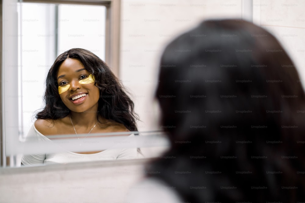 Parches para los ojos. Atractiva mujer africana de piel oscura en el baño con parches dorados transparentes en la piel debajo de los ojos, mirándose en el espejo y sonriendo. Higiene, concepto de cuidado facial.