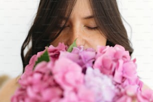 白い壁の背景に美しいアジサイの花束の匂いを嗅ぐ若い女性。ピンクと紫のアジサイの花を持っているおしゃれな女の子。母の日または女性の日おめでとう。