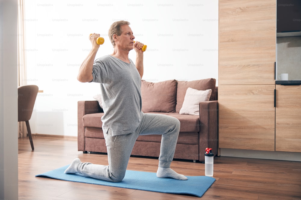 Kind adult man leaning knee on the mat while lifting dumbbells, making sporty body in room