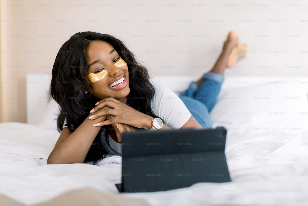 Relax and beauty procedures on weekend. Pleasant African smiling woman lying on bed in cozy room, with golden eye patches under her eyes and watching a film on ipad tablet or having a video call.