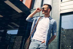 Handsome brunette expressing positivity while enjoying sunny urban day, wearing stylish sunglasses