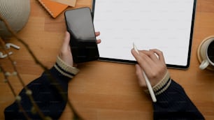 Overhead shot of female freelancer left hand holding smartphone and right hand working with mock-up tablet on wooden table
