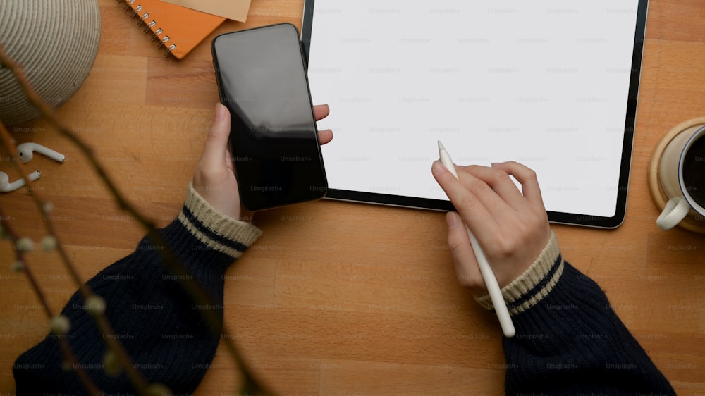 Overhead shot of female freelancer left hand holding smartphone and right hand working with mock-up tablet on wooden table