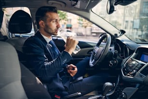Kind bearded man wearing official costume and sitting in his car, drinking fresh water