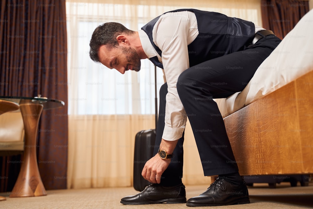 Low angle of cheerful young executive in elegant suit tying shoelaces while arriving with suitcase