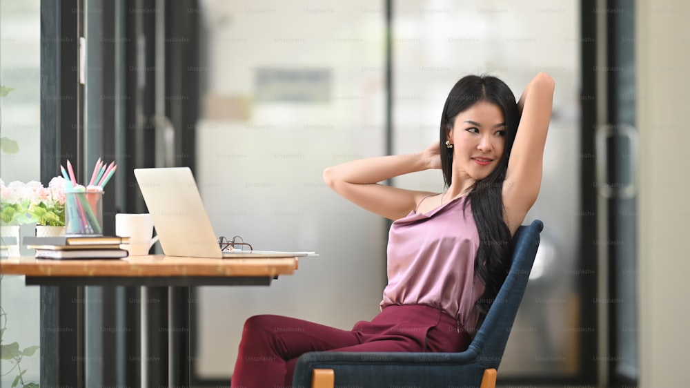 Asian woman is stretching her arms while sitting in front of a computer laptop at the wooden working desk.