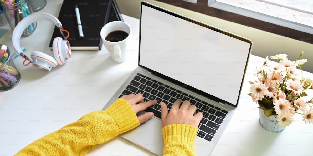 Cropped image of a woman's hands is typing on a white blank screen computer laptop at a white table.
