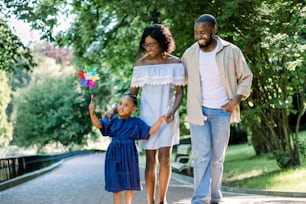 Happy family, joyful time together outdoors. Dark skinned little girl in blue dress, blowing colorful windmill toy, while walking in park alley with her young happy parents on summer day.