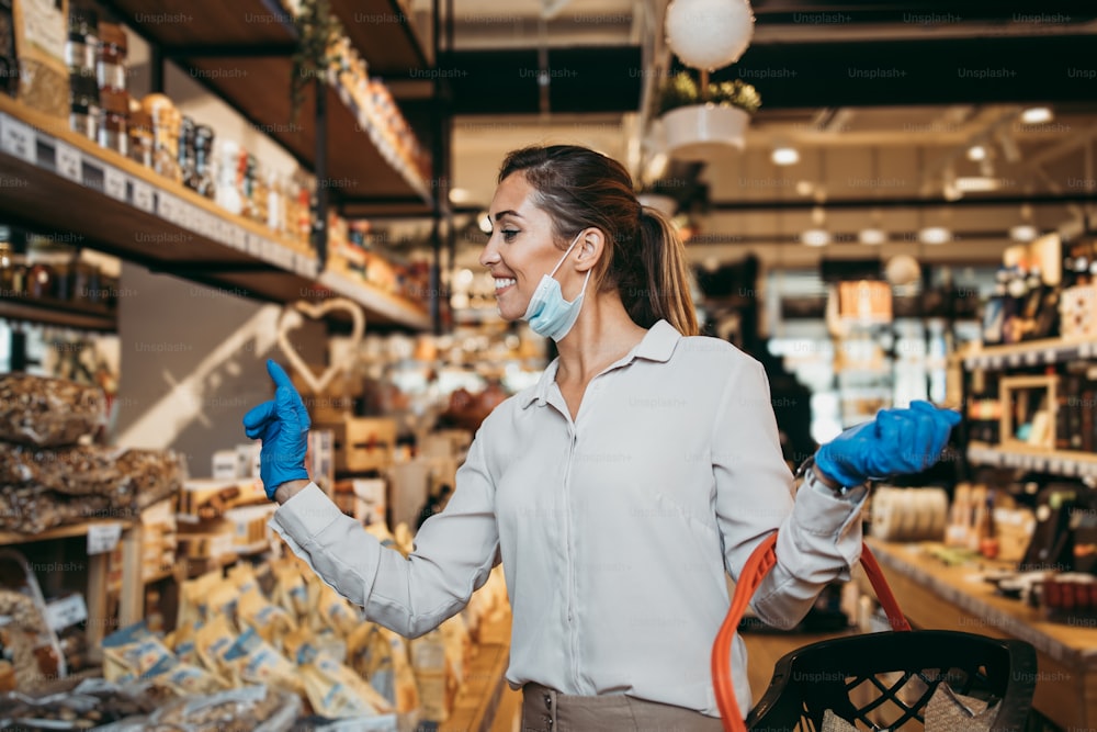 Beautiful young and elegant woman with face protective mask and gloves buying healthy food and drink in a modern supermarket or grocery store. Pandemic or epidemic lifestyle and consumerism concept.