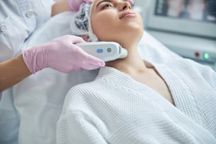 Experienced beautician performing neck lifting with modern equipment while calm patient lying on medical couch