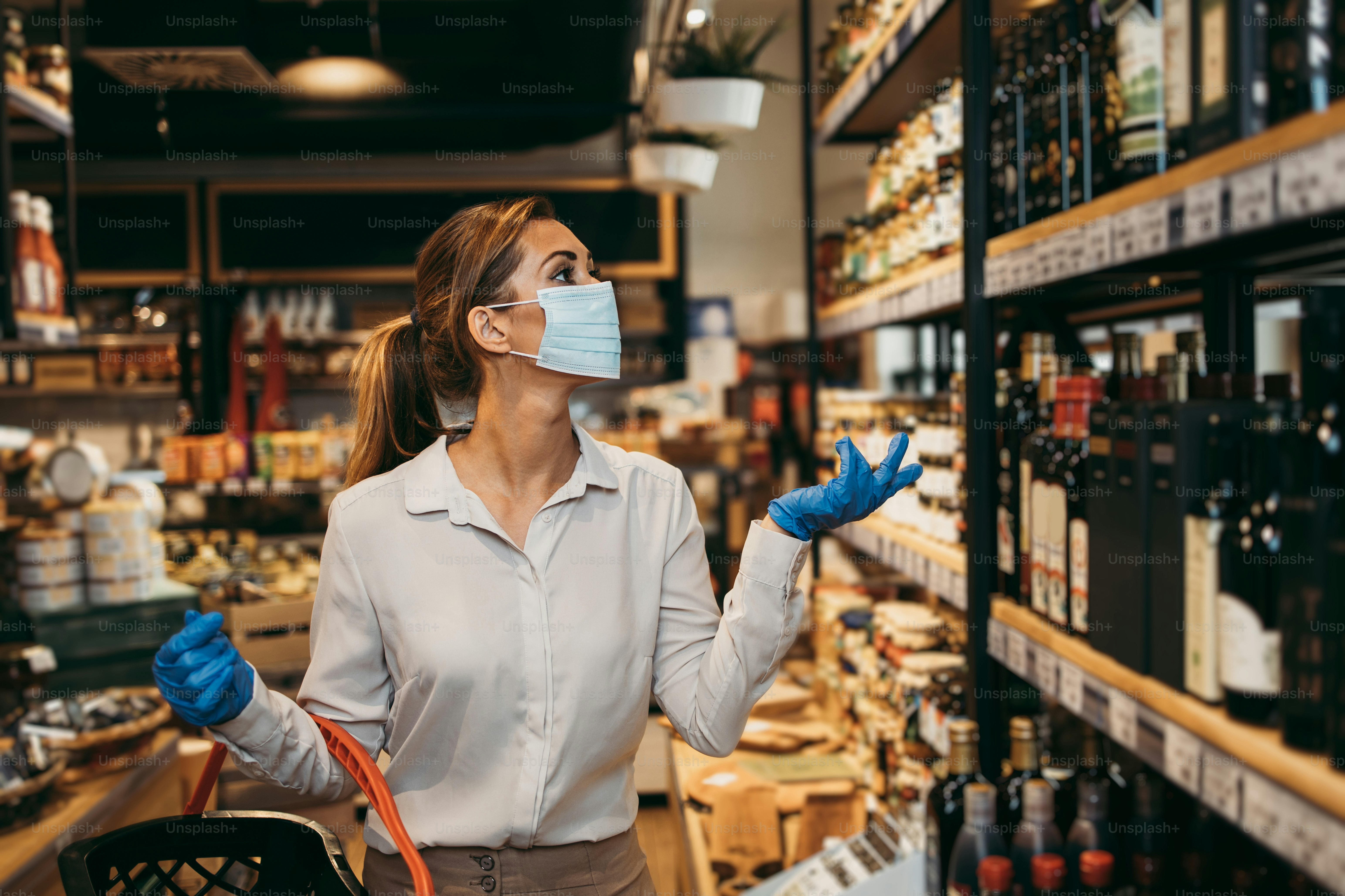 Beautiful young and elegant woman with face protective mask and gloves buying healthy food and drink in a modern supermarket or grocery store. Pandemic or epidemic lifestyle and consumerism concept.