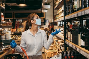 Beautiful young and elegant woman with face protective mask and gloves buying healthy food and drink in a modern supermarket or grocery store. Pandemic or epidemic lifestyle and consumerism concept.