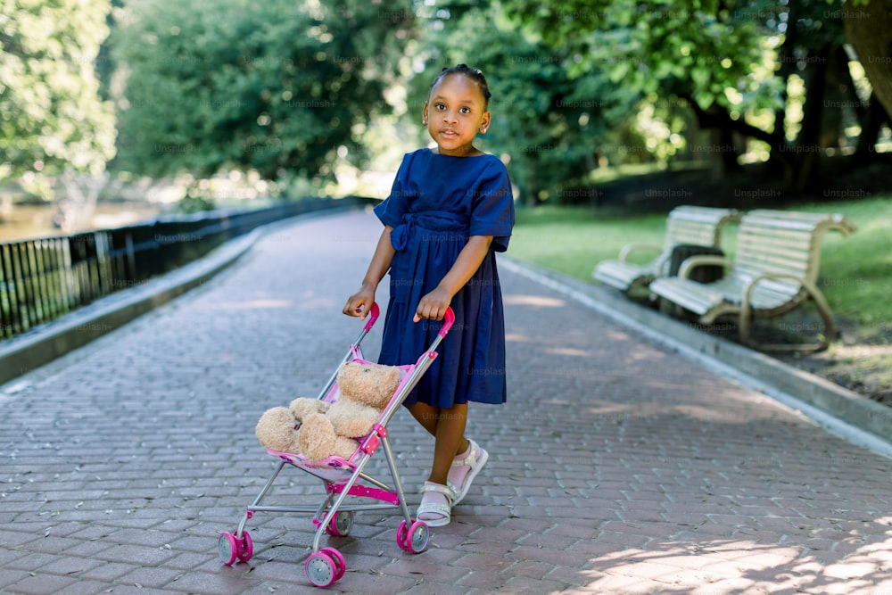 Kleines 5-jähriges afrikanisches Mädchen geht und spielt mit ihrem Spielzeugkinderwagen im Park. Süßes kleines dunkelhäutiges Baby mit Kinderwagen und Teddybär darin, das in einem Park posiert. Konzept der glücklichen Kindheit.