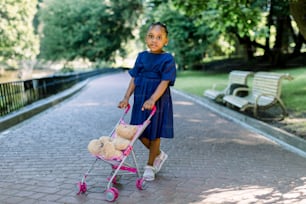 Little 5 years old African kid girl walk and playing with her toy stroller in park. Cute little dark skinned baby girl with a pram and teddy bear in it, posing in a park. Happy childhood concept.