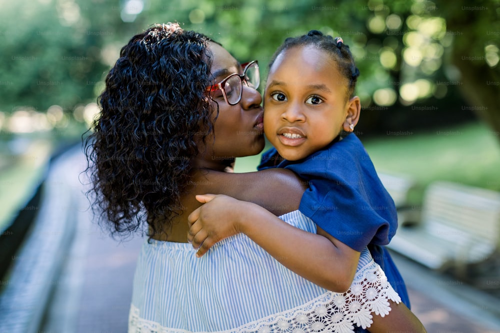 Close up portrait of little cute smiling African child girl in blue dress, looking at camera, while hugging pretty mother, kissing her in the cheek. African mother and daughter together in park.