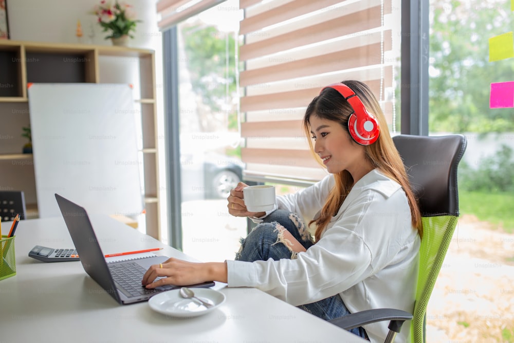 Retrato de una joven asiática que escucha música con auriculares y usa una computadora portátil y bebe café en la oficina.